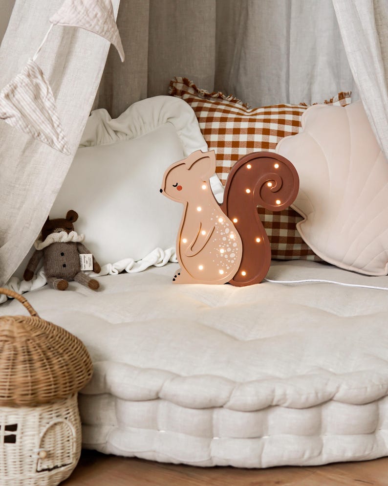 May include: A light-up squirrel-shaped night light in shades of tan and brown, with a curly tail. The lamp is on a white quilted surface, with pillows and a decorative basket in the background. The lamp is plugged in.