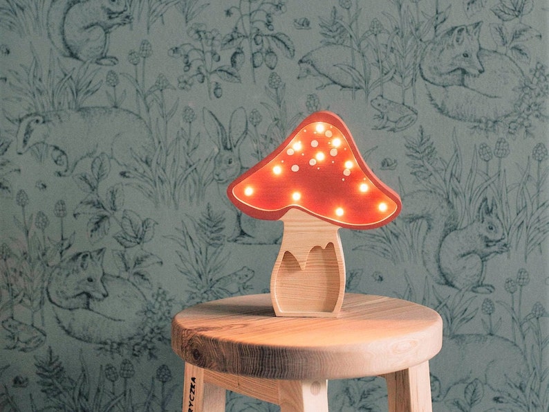 May include: A mushroom-shaped lamp with a light wood base and a red cap featuring small, circular cutouts that emit a warm, glowing light. The lamp is constructed from wood and is designed to stand upright on a flat surface, showcasing a natural grain.