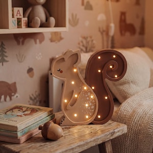 Puede incluir: Una luz de noche de madera con forma de ardilla con luces LED blancas cálidas. La luz está sobre una mesa de madera rústica, con libros y una bellota de madera. Un oso de peluche y bloques de alfabeto están en el fondo.