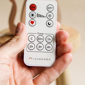 May include: A white remote control with black and red buttons and icons, including "ON", "OFF", a sun, a heart, and a moon. The remote has timer settings and the text "room GAGA".