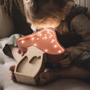 May include: A mushroom-shaped wooden lamp with a smooth, terracotta-colored cap featuring integrated warm LED lights. The base is crafted from light-toned natural wood with a visible grain, showcasing a cutout design on the front stem.