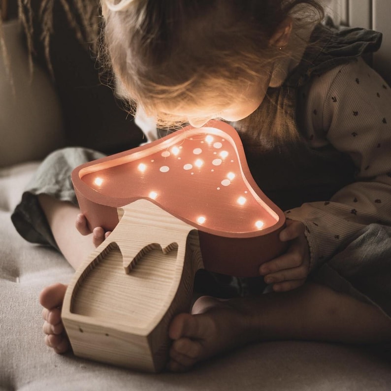 May include: A mushroom-shaped wooden lamp with a smooth, terracotta-colored cap featuring integrated warm LED lights. The base is crafted from light-toned natural wood with a visible grain, showcasing a cutout design on the front stem.