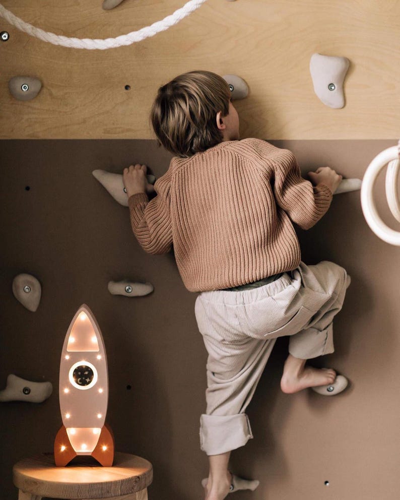 Op de afbeelding: Een decoratieve houten raketlamp met een warme gloed, met een witte behuizing en oranje accenten, staat op een kleine kruk. Een kind klimt op een klimmuur met beige grepen, gekleed in een beige trui en broek.