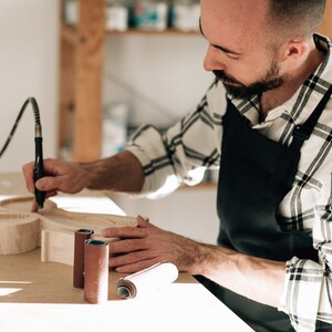 May include: A light-colored wooden object with rounded cutouts is being carved with a black rotary tool. Two cylindrical sanding rolls are placed on the work surface. The artisan wears a black apron over a white and black plaid long-sleeve shirt.