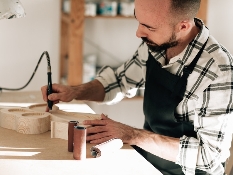 May include: A light-colored wooden object with rounded cutouts is being carved with a black rotary tool. Two cylindrical sanding rolls are placed on the work surface. The artisan wears a black apron over a white and black plaid long-sleeve shirt.