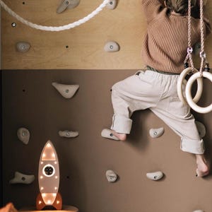 Op de afbeelding: Een kind klimt op een houten klimmuur met grijze klimgrepen. Een wit touw en houten gymnastiekringen hangen van bovenaf. Een verlichte raketvormige lamp met oranje accenten staat op een kleine houten kruk.