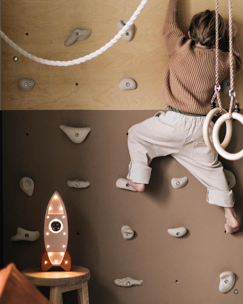 Op de afbeelding: Een kind klimt op een houten klimmuur met grijze klimgrepen. Een wit touw en houten gymnastiekringen hangen van bovenaf. Een verlichte raketvormige lamp met oranje accenten staat op een kleine houten kruk.