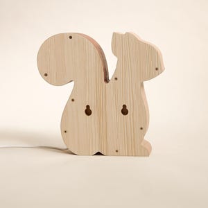 May include: Wooden squirrel-shaped night light. The light is made of light-colored wood and features a large, rounded tail and a small head. It has two holes for hanging and several screw heads are visible.