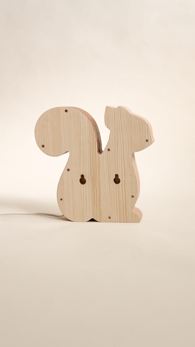 May include: Wooden squirrel-shaped night light. The light is made of light-colored wood and features a large, rounded tail and a small head. It has two holes for hanging and several screw heads are visible.