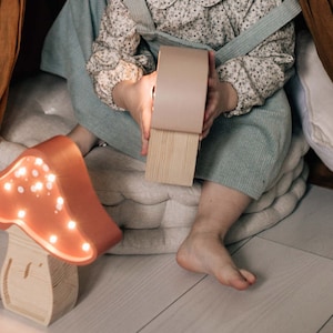 May include: A mushroom-shaped lamp with a terracotta-colored cap featuring small, glowing LED lights and a light-colored wooden base with visible grain. The lamp emits a warm, soft light, highlighting the smooth texture of the cap and the natural wood.