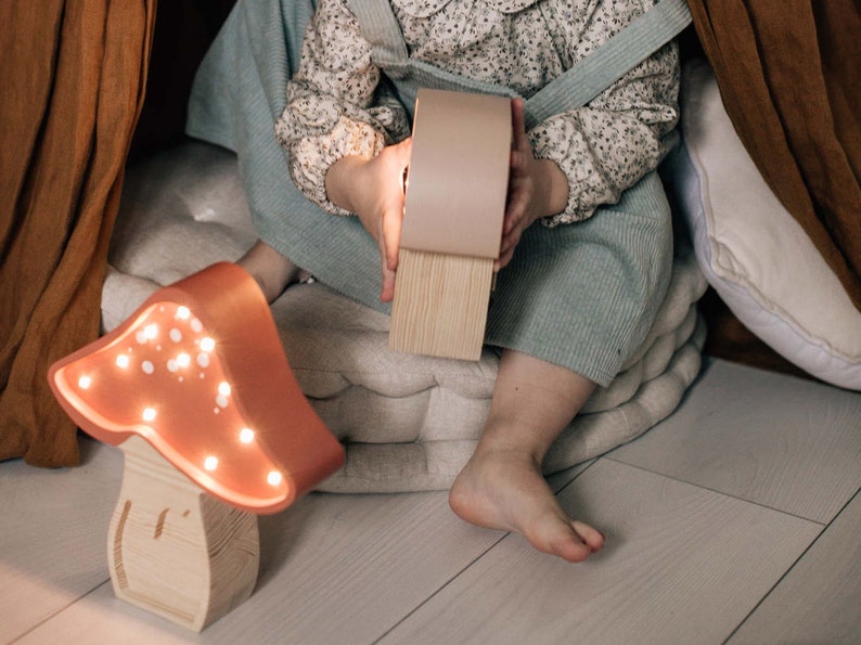 May include: A mushroom-shaped lamp with a terracotta-colored cap featuring small, glowing LED lights and a light-colored wooden base with visible grain. The lamp emits a warm, soft light, highlighting the smooth texture of the cap and the natural wood.