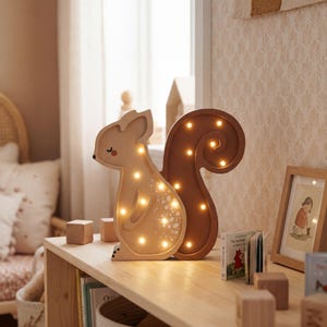May include: A wooden squirrel-shaped night light with warm white LED lights. The light has a light brown body and a darker brown tail. It sits on a wooden shelf with books and wooden blocks, creating a cozy nursery decor.
