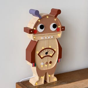 May include: A wooden robot-shaped night light with a light brown body, brown accents, and a purple antenna. The robot has large, cartoonish eyes, a meter on its chest, and small lights throughout. The night light is plugged into a wall outlet.