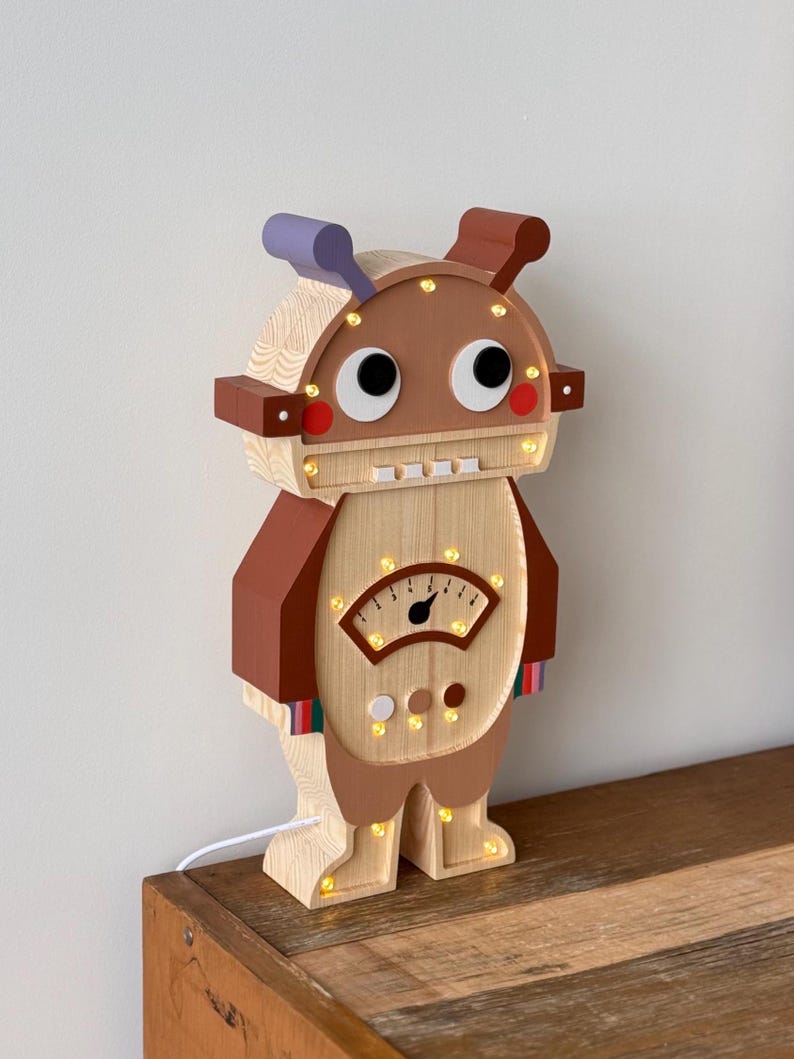 May include: A wooden robot-shaped night light with a light brown body, brown accents, and a purple antenna. The robot has large, cartoonish eyes, a meter on its chest, and small lights throughout. The night light is plugged into a wall outlet.