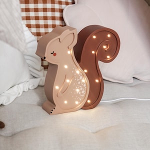 May include: A light-up squirrel-shaped night light in shades of brown and tan. The squirrel has a curved tail and a series of small lights that illuminate the body. The night light is sitting on a bed with pillows in the background.