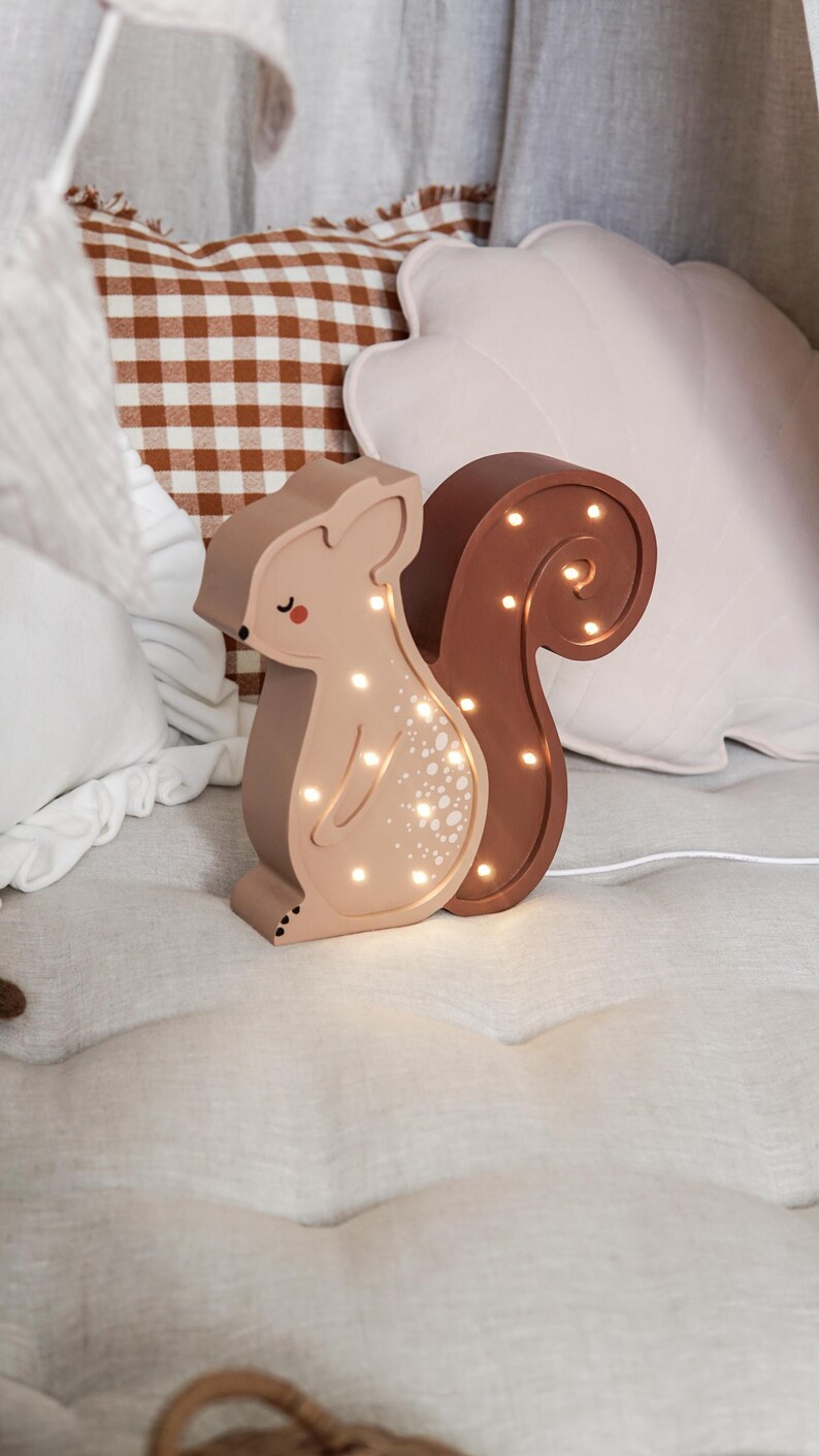 May include: A light-up squirrel-shaped night light in shades of brown and tan. The squirrel has a curved tail and a series of small lights that illuminate the body. The night light is sitting on a bed with pillows in the background.