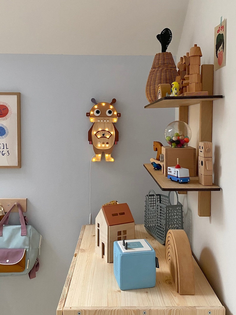 May include: A wooden robot-shaped wall light with warm LED lights is mounted on a light blue wall. A wooden house, a blue speaker, and other wooden toys sit on a wooden table. A backpack hangs on the wall to the left.