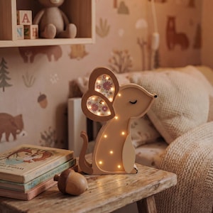 May include: A wooden mouse-shaped night light with illuminated dots and floral-patterned ears. The light sits on a rustic wooden table next to books and a wooden acorn. A teddy bear and alphabet blocks are on a shelf in the background.