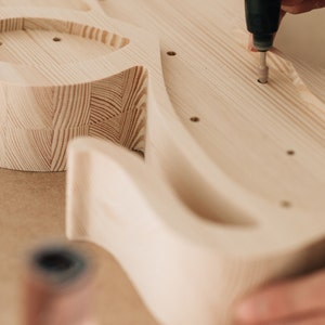 May include: A light-colored wooden guitar body being sanded with a rotary tool. The unfinished wood shows a visible grain pattern with several pre-drilled holes. The tool is positioned over one of the holes, refining the surface of the instrument.