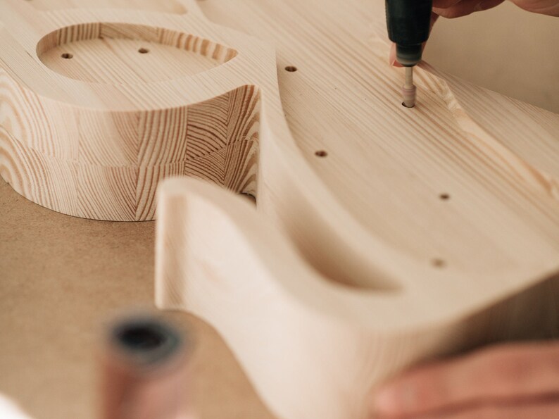May include: A light-colored wooden guitar body being sanded with a rotary tool. The unfinished wood shows a visible grain pattern with several pre-drilled holes. The tool is positioned over one of the holes, refining the surface of the instrument.