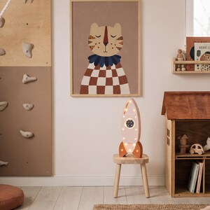 Op de afbeelding: Een kinderkamersc&egrave;ne met een ingelijste tijgerillustratie met een bruin en wit geblokt patroon. Een roze raketvormige lamp met verlichting staat op een kleine houten kruk. Een houten klimmuur en poppenhuis zijn ook zichtbaar.