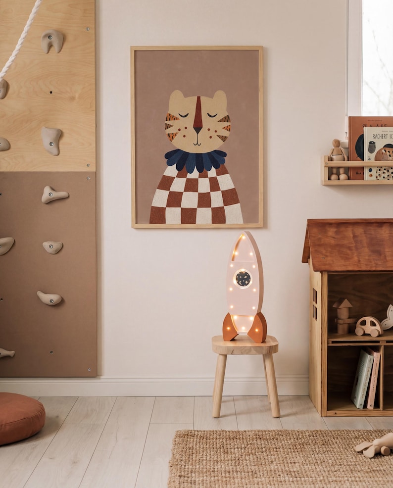 Op de afbeelding: Een kinderkamersc&egrave;ne met een ingelijste tijgerillustratie met een bruin en wit geblokt patroon. Een roze raketvormige lamp met verlichting staat op een kleine houten kruk. Een houten klimmuur en poppenhuis zijn ook zichtbaar.