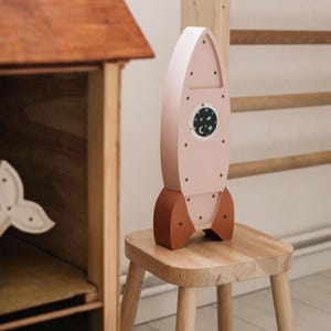 Op de afbeelding: Een houten speelgoedraket in lichtroze met bruine accenten. De raket heeft een rond venster met een nachthemelontwerp en staat op een kleine houten kruk. Het speelgoed is voor kinderen.