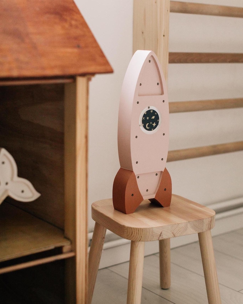 Op de afbeelding: Een houten speelgoedraket in lichtroze met bruine accenten. De raket heeft een rond venster met een nachthemelontwerp en staat op een kleine houten kruk. Het speelgoed is voor kinderen.