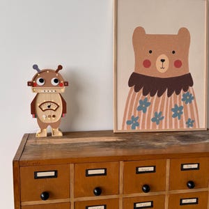 May include: A wooden robot-shaped lamp with a light-up dial and a framed bear illustration. The bear illustration features a brown bear wearing a floral shirt. The lamp and illustration are on a wooden cabinet with multiple drawers.