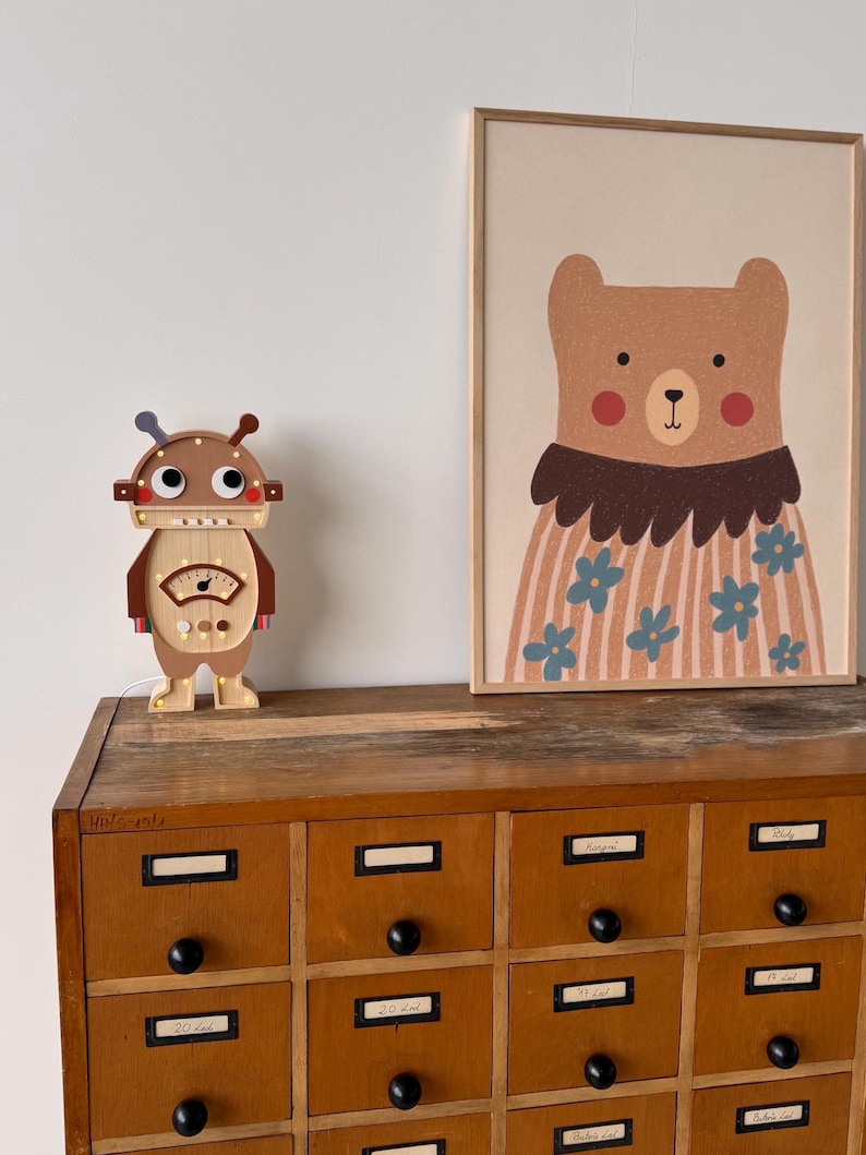 May include: A wooden robot-shaped lamp with a light-up dial and a framed bear illustration. The bear illustration features a brown bear wearing a floral shirt. The lamp and illustration are on a wooden cabinet with multiple drawers.