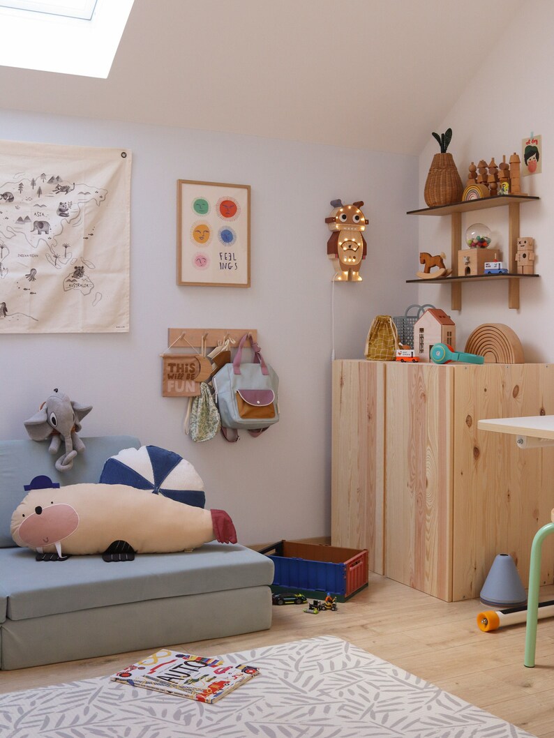 May include: A child's room with a light blue sofa, a walrus-shaped pillow, and a wooden cabinet. The walls are decorated with a world map, a framed artwork, and a wooden light fixture. Toys and decorative items are on shelves.