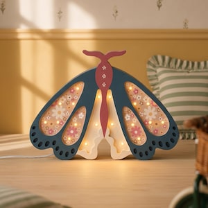 May include: A butterfly-shaped lamp with a dark blue frame and a pink central body. The wings feature cutouts revealing a light-colored background patterned with small pink and white flowers, illuminated by several small warm-toned LED lights.
