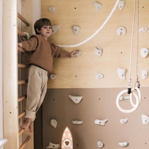 Op de afbeelding: Een indoor klimmuur met een houten ladder, klimgrepen en een touw. Een raketvormige lamp met een warme gloed staat eronder. Witte gymnastiekringen hangen aan het plafond.