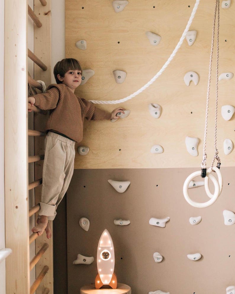 Op de afbeelding: Een indoor klimmuur met een houten ladder, klimgrepen en een touw. Een raketvormige lamp met een warme gloed staat eronder. Witte gymnastiekringen hangen aan het plafond.