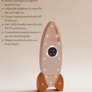 Op de afbeelding: Een roze houten raket nachtlampje met warme LED-verlichting. De raket heeft bruine vinnen en een rond raam met een sterrenhemel ontwerp. De tekst zegt "Handcrafted from solid pine wood - durable & eco-friendly".