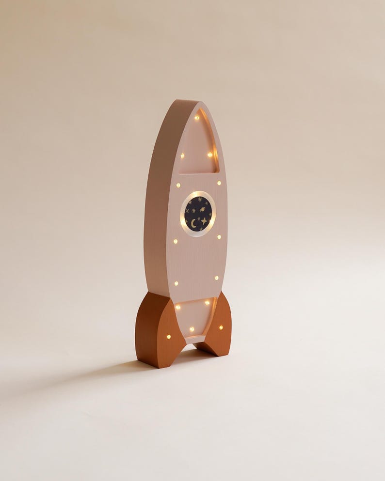 Op de afbeelding: Houten nachtlampje in de vorm van een raket, roze en bruin. De raket heeft een rond venster met een sterrenhemelontwerp en kleine lampjes. De basis en vinnen zijn bruin, de body is roze. Ongeveer 30 cm hoog.