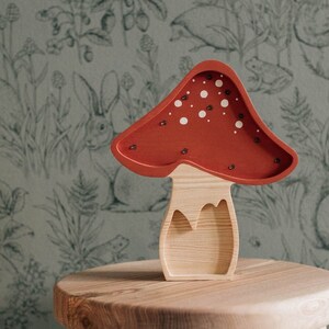 May include: A wooden mushroom-shaped lamp with a red cap featuring white circular spots and small embedded lights. The stem is made of light-colored natural wood with a visible grain, and the overall structure is hollowed out to form a decorative piece.