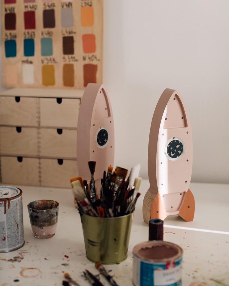 Op de afbeelding: Twee roze houten raket speelgoed met oranje vinnen en een cirkelvormig raam met een nachthemel ontwerp. Een verzameling penselen staat in een groene metalen container, met verfblikken en een houten kleurenkaart op de achtergrond.