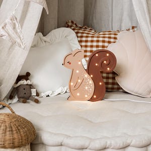May include: A light-up squirrel-shaped night light with a brown tail and a tan body. The night light is on a white, tufted surface, with a small stuffed animal and a woven basket nearby. The background includes pillows and draped fabric.