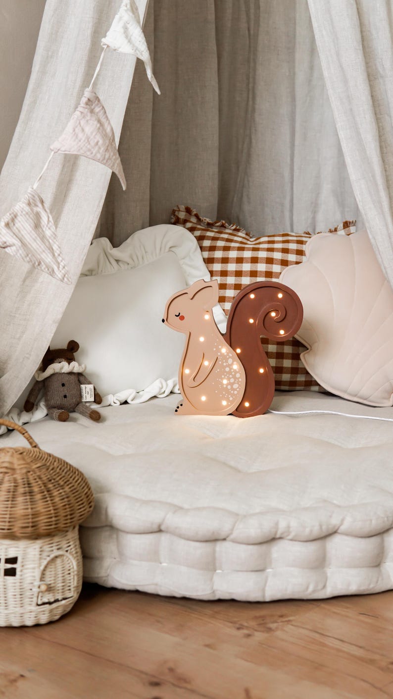 May include: A light-up squirrel-shaped night light with a brown tail and a tan body. The night light is on a white, tufted surface, with a small stuffed animal and a woven basket nearby. The background includes pillows and draped fabric.