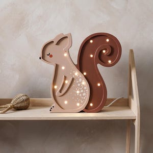 May include: A wooden squirrel-shaped night light in shades of brown and tan. The light has a curled tail and is illuminated with small, warm white lights. It sits on a wooden shelf with other toys.