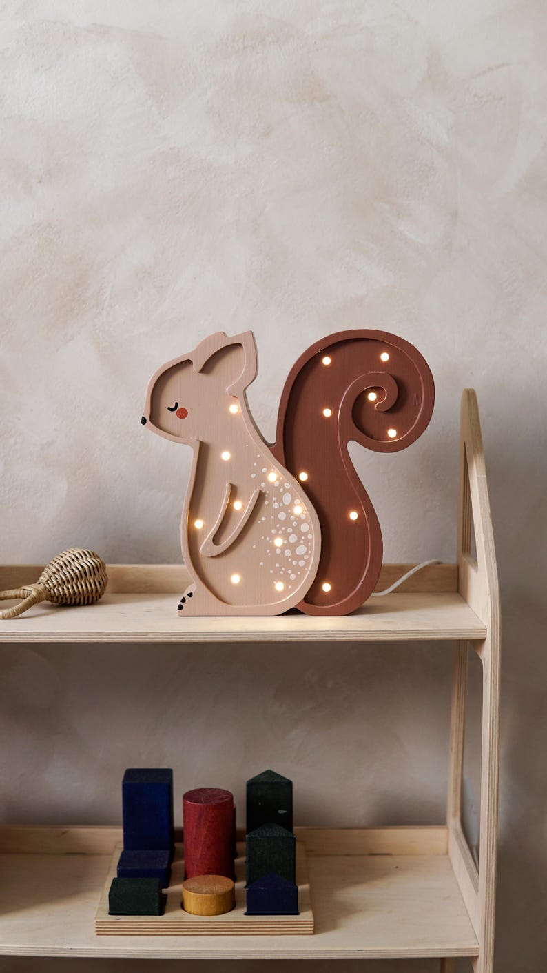 May include: A wooden squirrel-shaped night light in shades of brown and tan. The light has a curled tail and is illuminated with small, warm white lights. It sits on a wooden shelf with other toys.