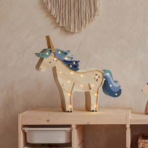 May include: Wooden unicorn night light with blue and teal accents, and star details. The unicorn has a light-colored body with small lights throughout. It is displayed on a wooden shelf with storage bins and a woven basket.