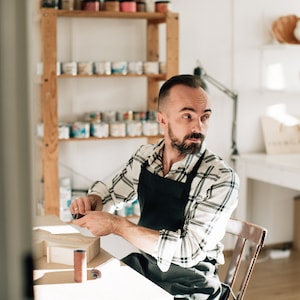 May include: A person wearing a black apron over a white and black plaid long-sleeved shirt, sitting at a wooden table while holding a small cylindrical sanding tool, with a wooden object and another sanding roll on the table surface.