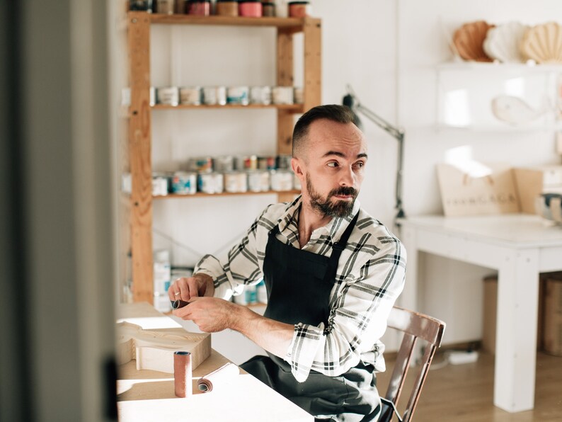 May include: A person wearing a black apron over a white and black plaid long-sleeved shirt, sitting at a wooden table while holding a small cylindrical sanding tool, with a wooden object and another sanding roll on the table surface.