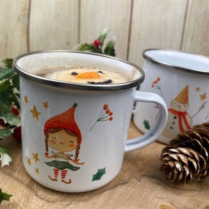 Puede incluir: Tazas de esmalte blanco con diseños de invierno. Una taza presenta un elfo con sombrero rojo y falda verde, la otra un muñeco de nieve. Las tazas contienen una bebida caliente con una cara de muñeco de nieve.