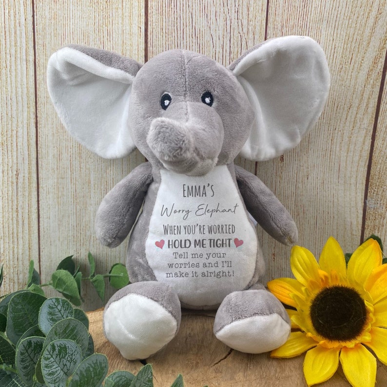 Op de afbeelding: Een grijze pluche olifant met grote oren en een slurf. De olifant heeft een witte vlek op zijn borst met de tekst "EMMA'S Worry Elephant" en een geruststellende boodschap. Een gele zonnebloem en groen zijn op de achtergrond.
