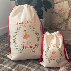 May include: Two beige drawstring gift bags with red drawstrings. The bags feature a watercolor design of a ballerina inside a wreath, with the text "Special Delivery For Anna". The bags are different sizes.