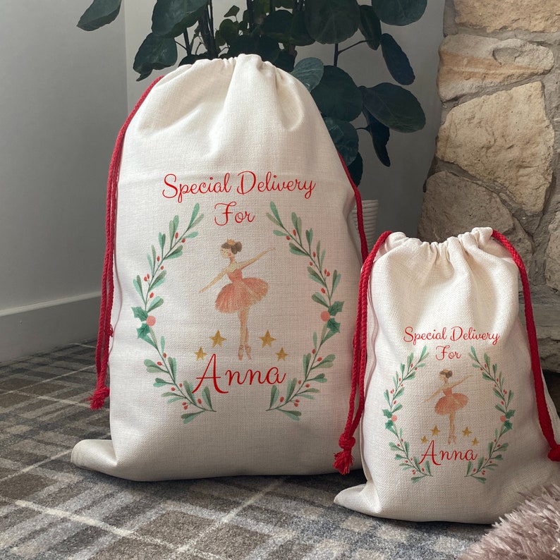 May include: Two off-white drawstring gift bags with red drawstrings. Each bag features a watercolor design of a ballerina inside a wreath with the text "Special Delivery For Anna". The bags are different sizes.