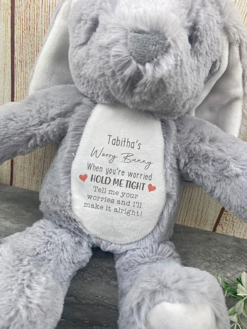 Op de afbeelding: Een grijze pluche konijn met lange oren. Het konijn heeft een witte patch op de borst met de tekst "Tabitha's Worry Bunny" en de zinnen "When you're worried HOLD ME TIGHT" en "Tell me your worries and I'll make it alright!"